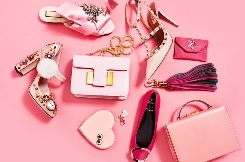 Various pink and beige ladies' accessories including high-heeled shoes, handbags, a wallet, a tassel keychain, and a heart-shaped pouch on a pink background.