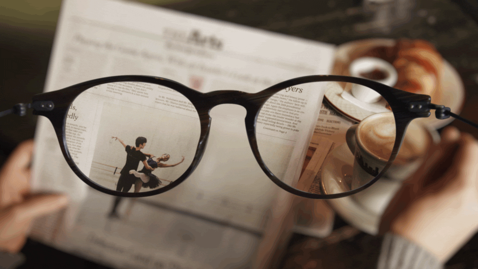 A pair of eyeglasses held over a newspaper featuring an image of two ballet dancers, with the focus through the lenses; a blurred cup of coffee is in the background.