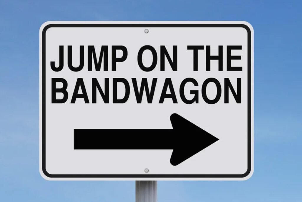 A traffic sign with the text "JUMP ON THE BANDWAGON" and a black arrow pointing to the right, against a clear blue sky.