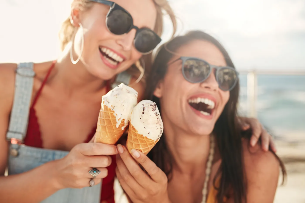 Two joyful women in sunglasses holding ice creams at the beach.