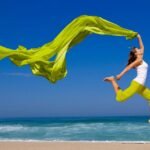 Woman joyfully leaps with flowing green fabric on tranquil beach under clear blue sky.
