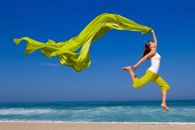 Woman joyfully leaps with flowing green fabric on tranquil beach under clear blue sky.