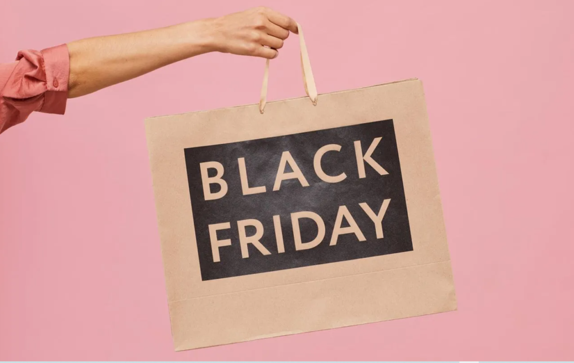 Adult arm holding Black Friday shopping bag on pink backdrop.