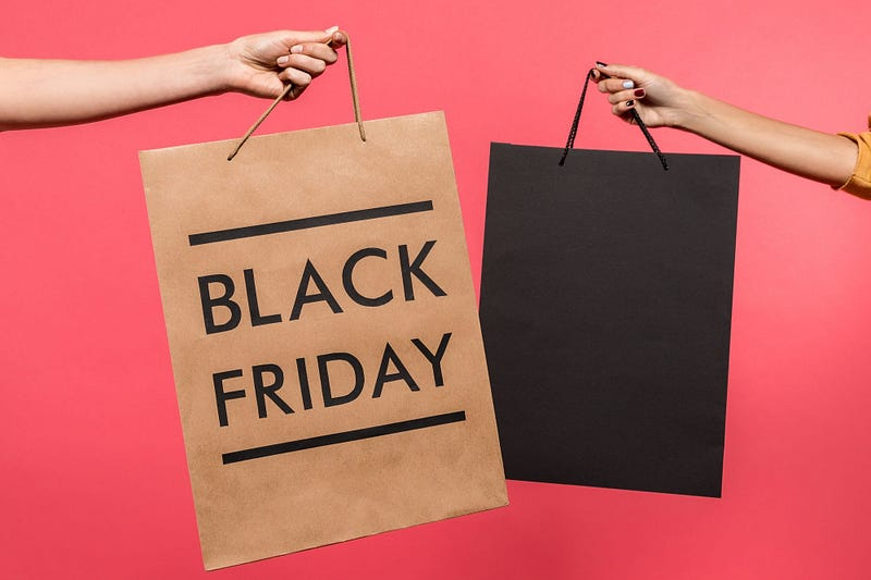 Two hands holding shopping bags against a pink background, one bag is brown with "BLACK FRIDAY" text and the other is plain black.