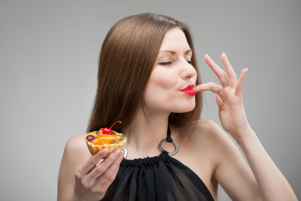 A woman with red lipstick and a black top blissfully tasting a fruit tart decorated with a cherry.