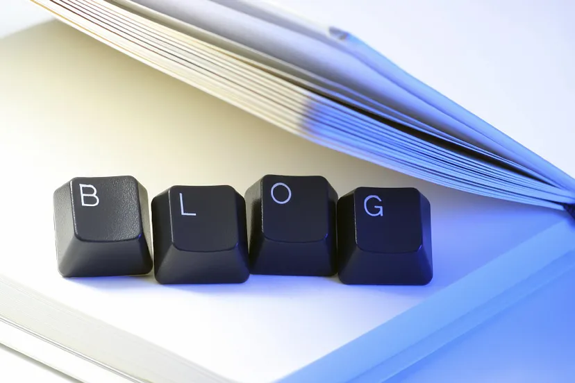 Four keyboard keys with letters B, L, O, G arranged to spell 'BLOG', resting on a white surface with an open book in the background.