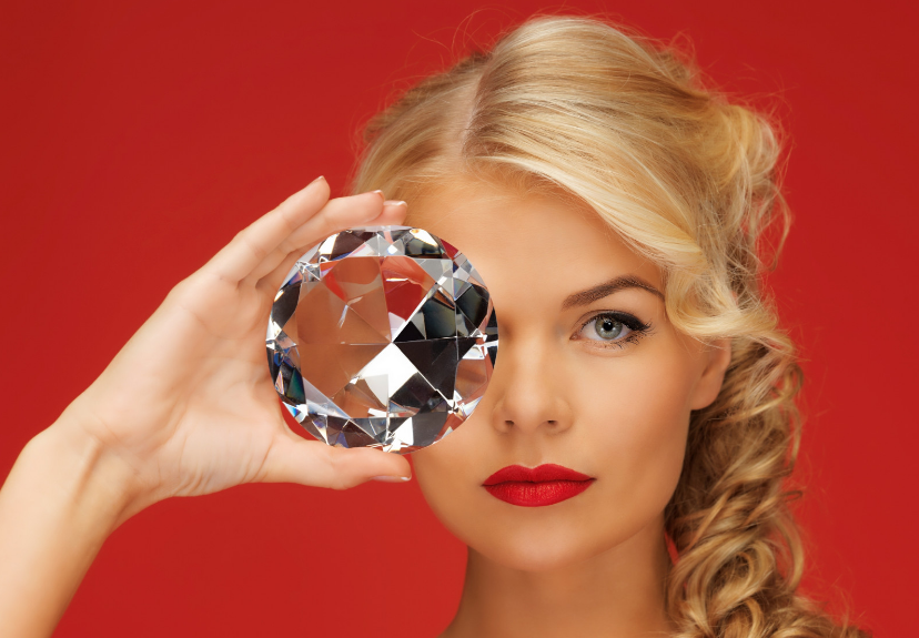 A woman with blonde hair and red lipstick holds a large, faceted gemstone in front of one eye, against a red background.