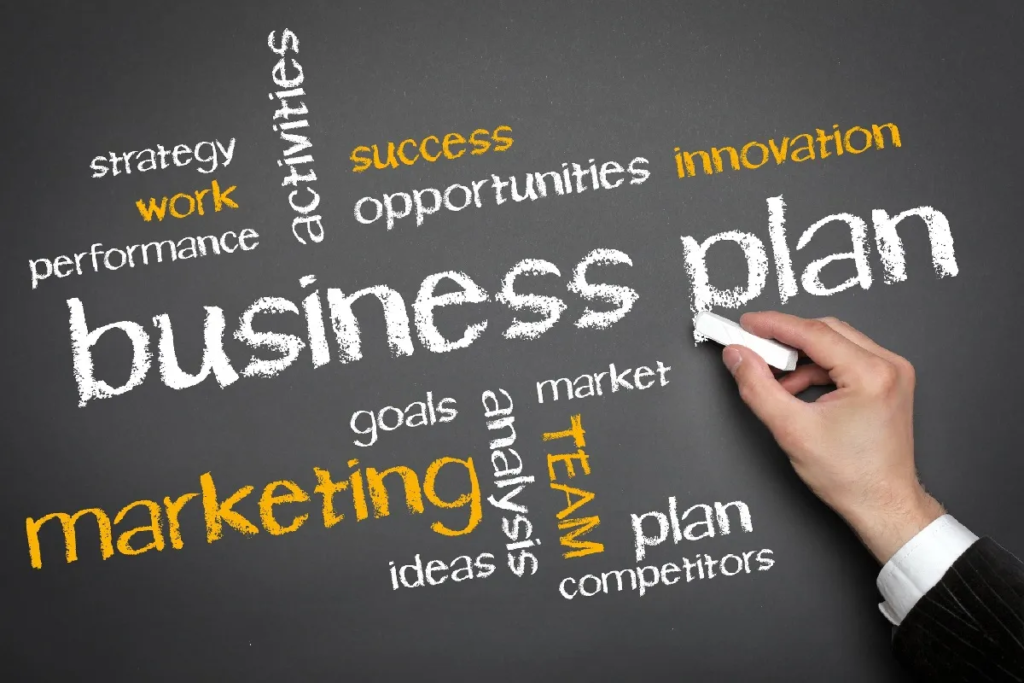 A person's hand writing the words "business plan" on a blackboard surrounded by related concepts such as strategy, marketing, goals, opportunities, and innovation written in various colours.