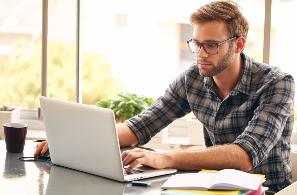 A man in glasses working intently on a laptop at a desk with a coffee cup and a notebook beside him, in a well-lit, modern interior.