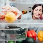 Cheerful woman selecting fresh fruits and veggies from refrigerator in a bright kitchen.