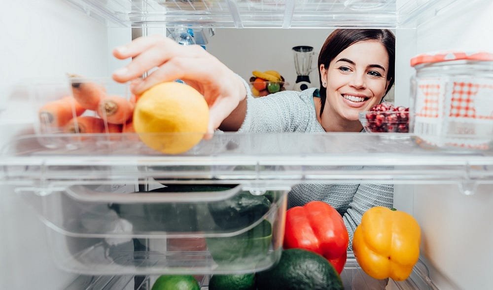 Cheerful woman selecting fresh fruits and veggies from refrigerator in a bright kitchen.