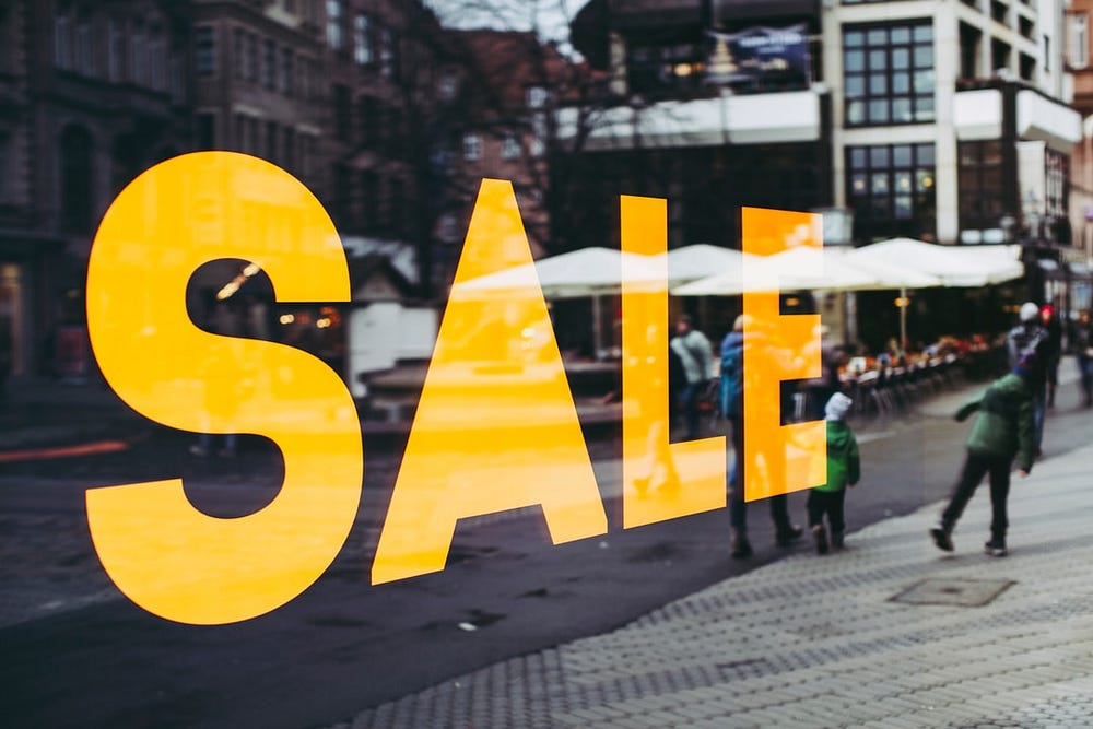 A storefront window with the word "SALE" displayed in large, bold, orange letters, slightly blurred people walking by on the street visible in the background.