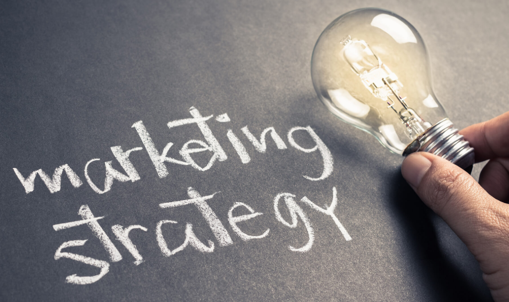 A hand holding a clear light bulb with "marketing strategy" written in chalk on a blackboard surface beneath it.