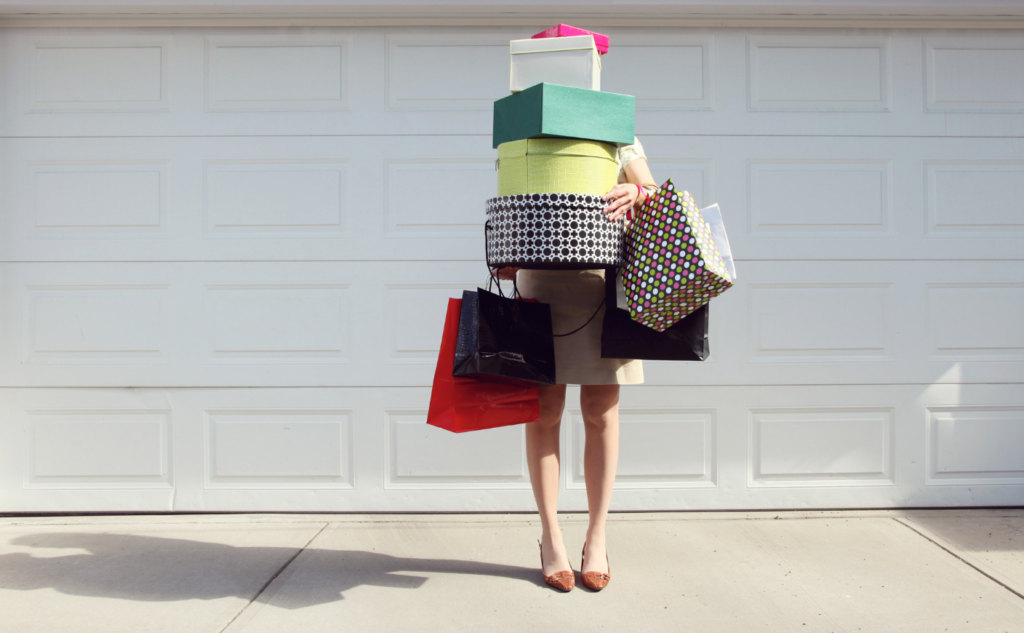 A person standing in front of a white garage door, holding a large stack of colorful gift boxes and shopping bags, obscuring their face and upper body.