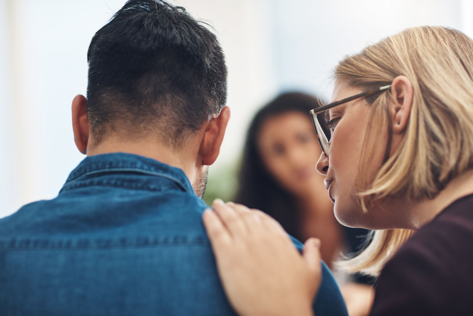 A close-up of a woman with glasses comforting a man by placing her hand on his shoulder, with an out-of-focus woman in the background.