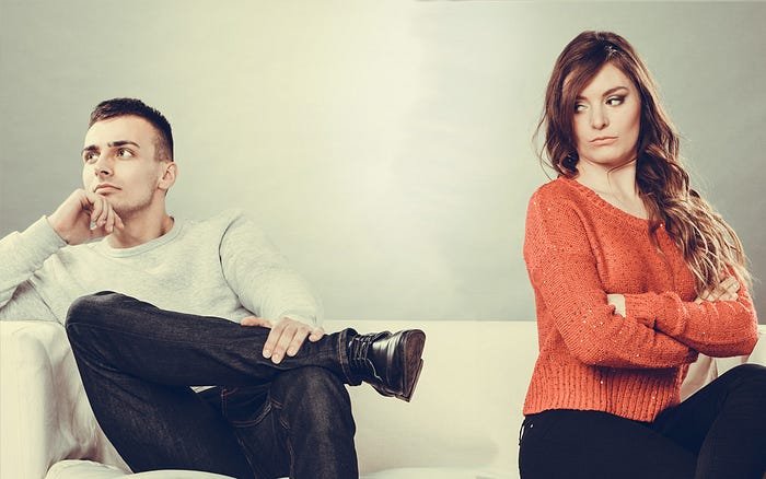 A man and a woman sitting at opposite ends of a couch, both looking away from each other with expressions of contemplation and displeasure.