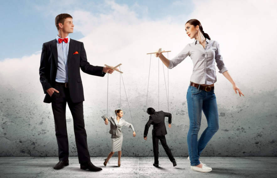 A man in a suit with a red bow tie and a woman in casual clothes are holding marionette controls connected to two business people posed as if they are puppets, set against a cloudy sky backdrop.