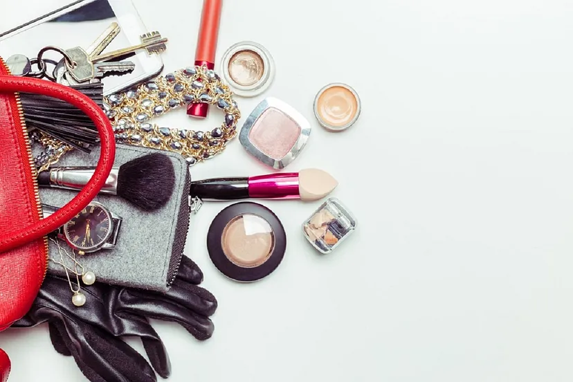 An assortment of cosmetic items and accessories, including makeup brushes, a red purse, eyeshadow, a necklace, a watch, black gloves, and keys, spread out on a white background.