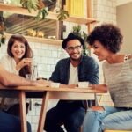 Friends enjoying coffee and laughter in trendy cafe with lush greenery and cozy ambiance.