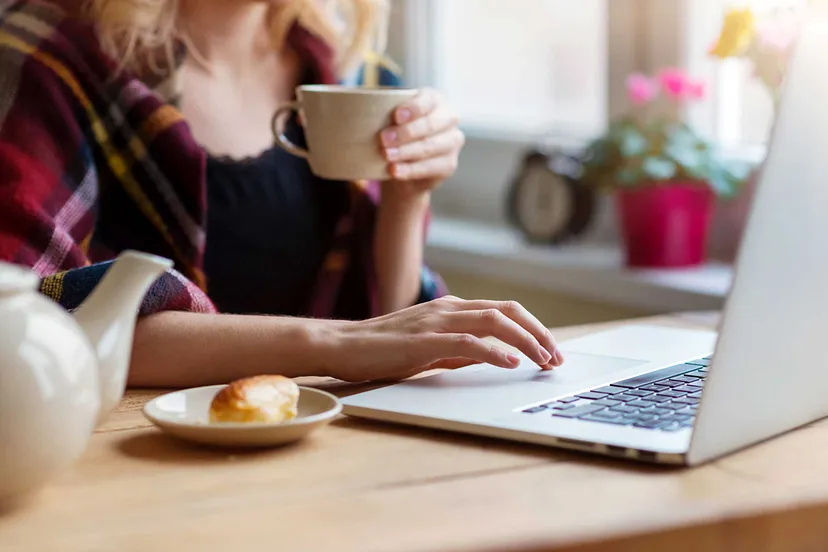 Woman holding a coffee cup and typing on a laptop with a pastry on a plate and a tea pot nearby, in a home environment.