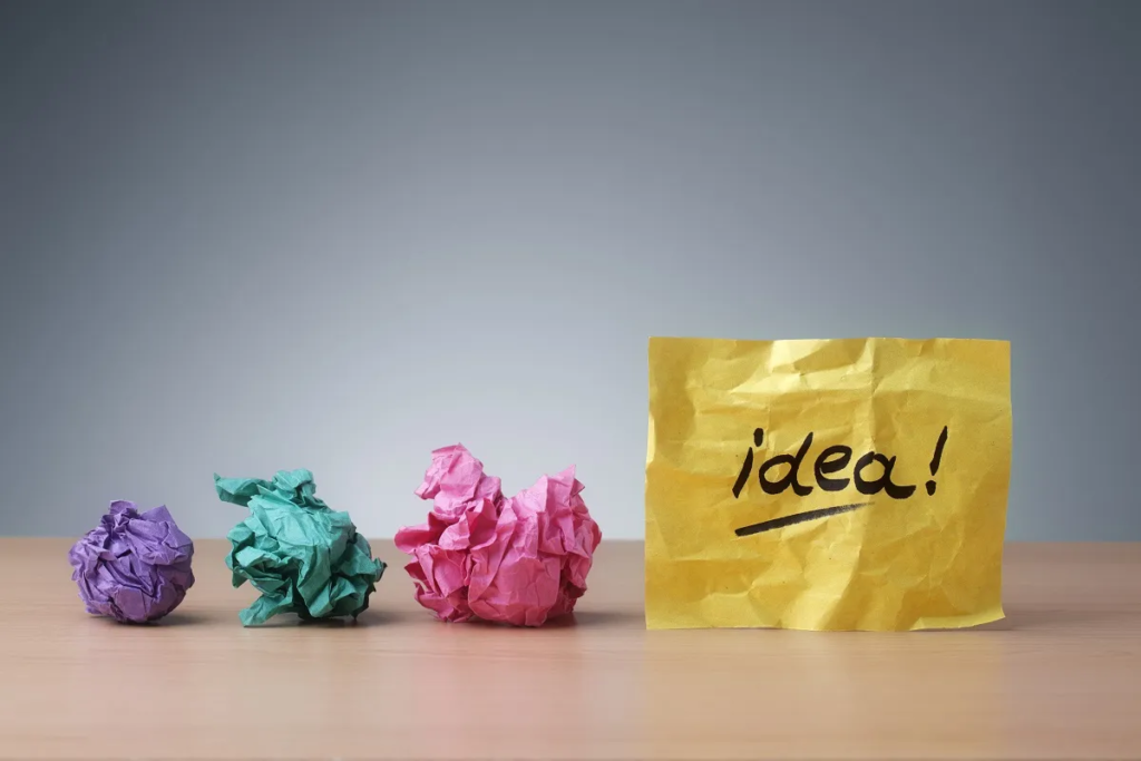 Crumpled balls of paper in blue, green, and pink on a wooden surface with a yellow piece of paper standing upright, having the word "idea!" handwritten on it, against a grey background.