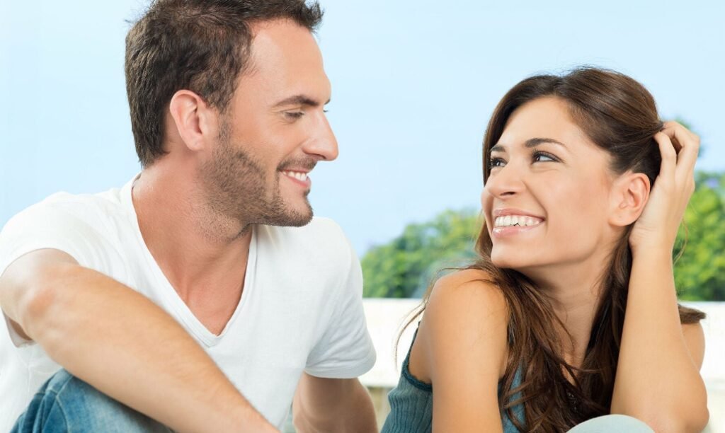 A man and a woman sitting closely, smiling at each other against a blue sky background with green foliage in the distance. They appear to be in a casual, joyful conversation.