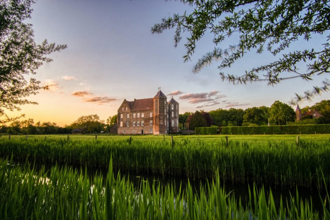 Tranquil manor house by green fields, pond, and sunset sky.