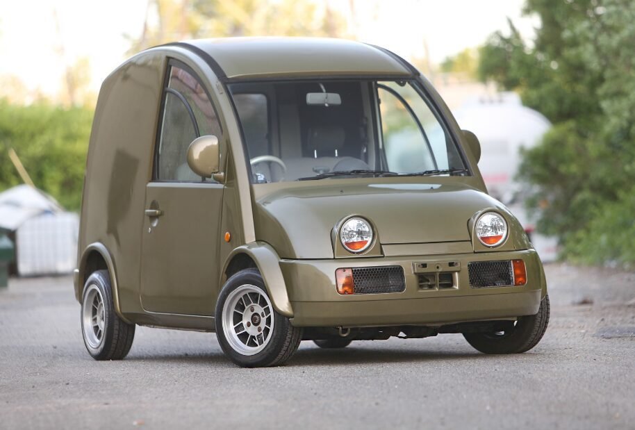 A small olive-green microcar parked on a street, featuring a distinctive tall and boxy rear design and large circular headlights.