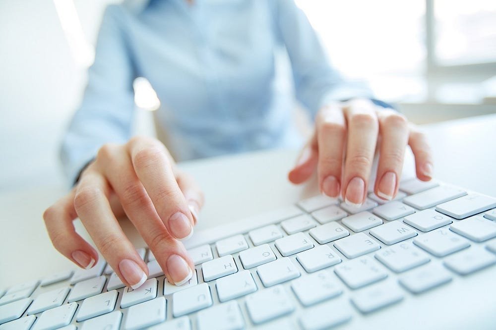 A close-up of a person's hands typing on a white computer keyboard, with a blurred background suggesting an office environment.