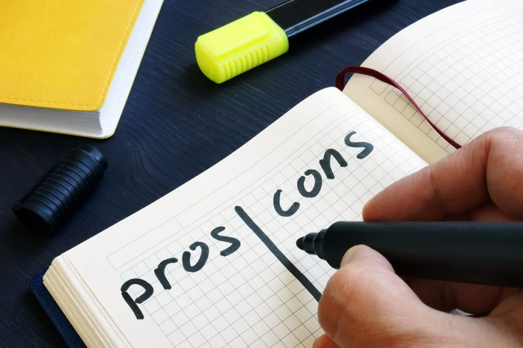 A person's hand writing a list with the headings "pros" and "cons" in a notebook, with a yellow highlighter and a black marker cap on the table.