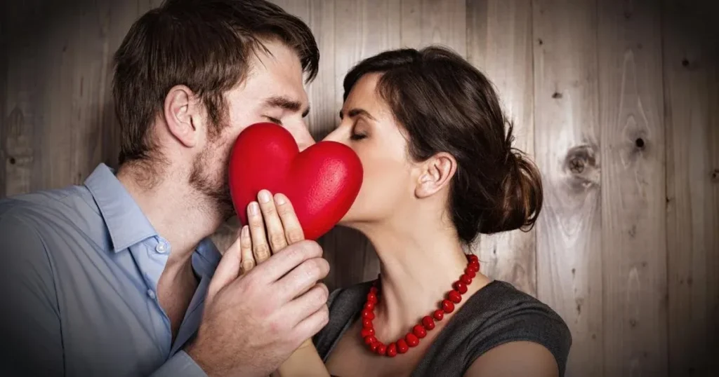 A man and a woman about to kiss, holding a red heart between them, with a rustic wooden background.