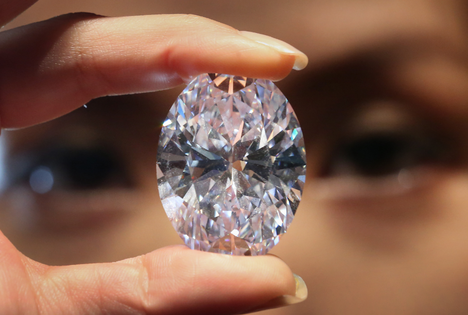 A close-up of a person's fingers holding a large, oval-cut diamond with the individual's out-of-focus eyes visible in the background.