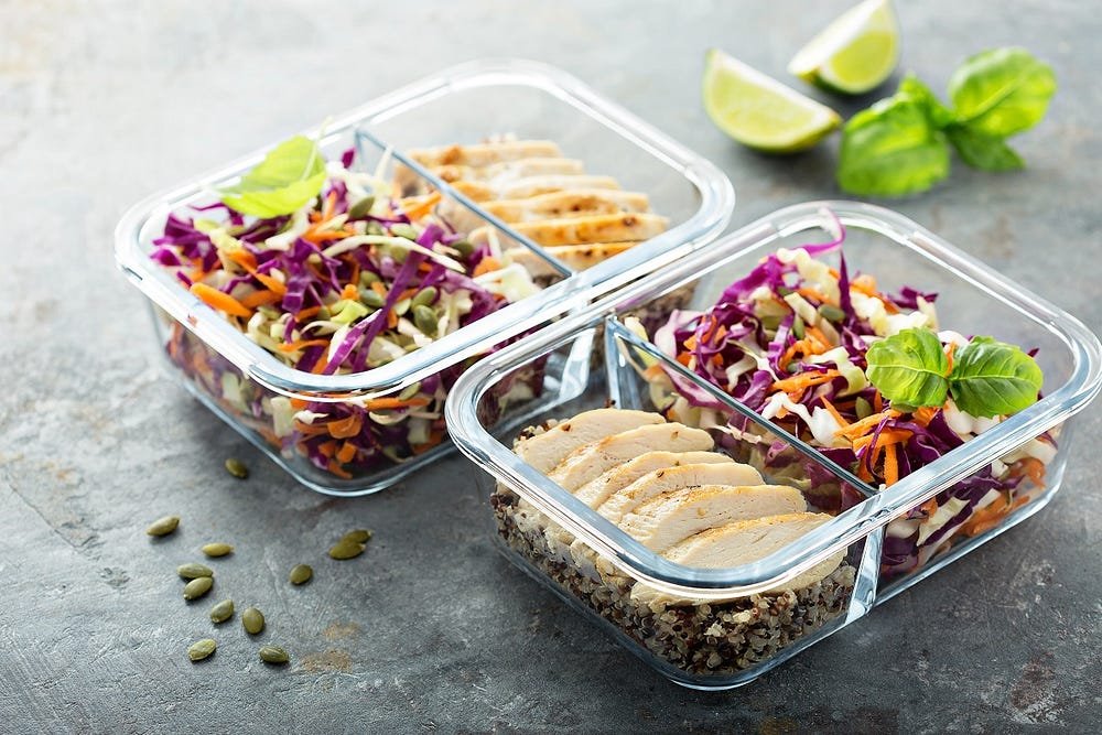 Three glass containers with quinoa, grilled chicken slices, and mixed vegetables including purple cabbage and carrots, garnished with basil leaves, with lime wedges and pumpkin seeds scattered on the side.