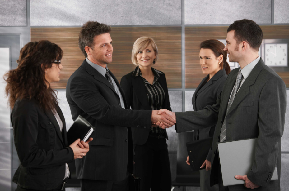 Two businessmen shaking hands in an office setting, with three female colleagues observing, one with a notebook and another with a file, all dressed in professional attire.