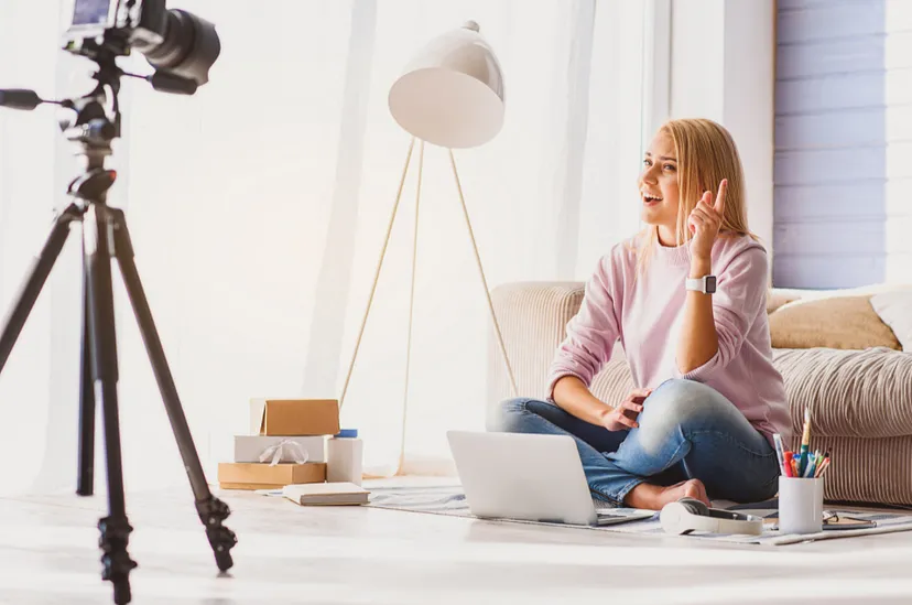 A woman sitting on the floor with a laptop and small boxes, waving at a camera on a tripod, possibly recording a vlog or engaging in a video call.