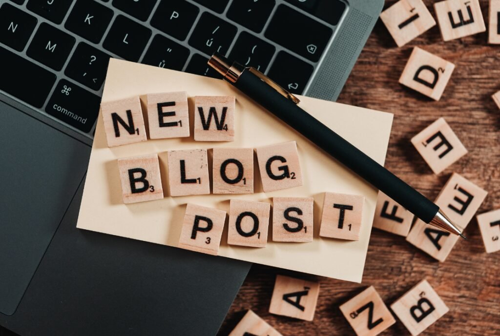 A laptop keyboard with a note on top displaying Scrabble tiles spelling "NEW BLOG POST" and a pen resting across the note, with more Scrabble tiles scattered around on a wooden surface.