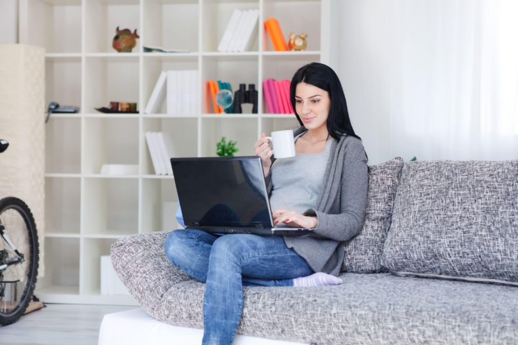 A woman sitting on a grey sofa with a laptop on her lap, holding a white mug, with a bookshelf in the background.