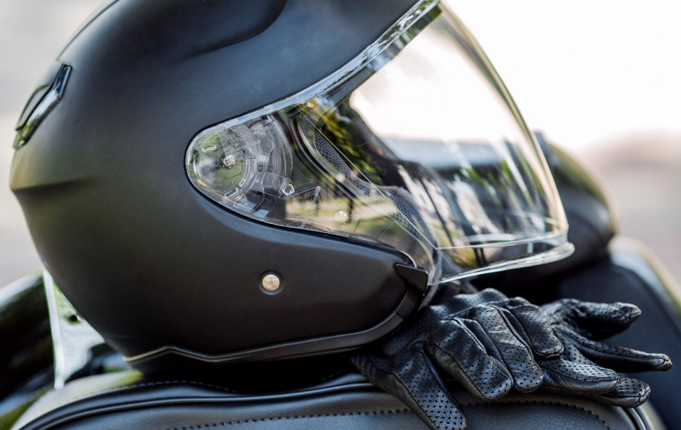 A close-up of a matte black motorcycle helmet with its visor partially open, resting on a seat next to a pair of black leather motorcycle gloves.