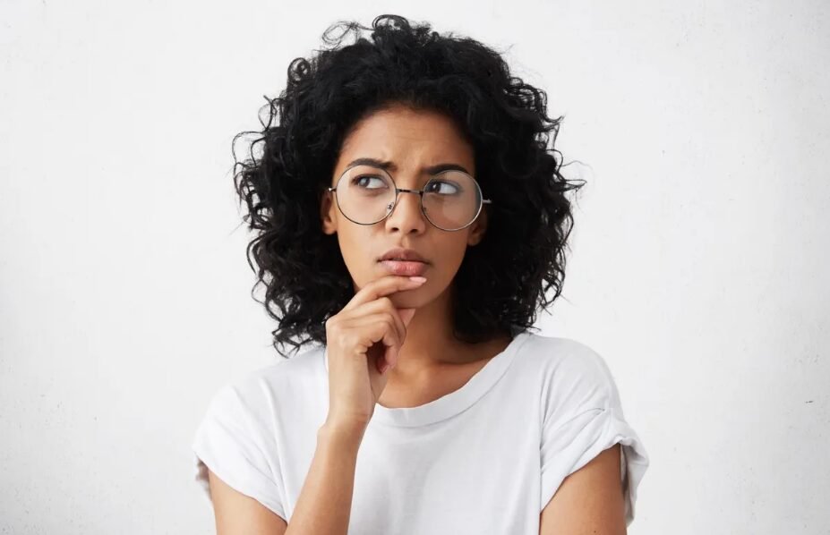Contemplative young woman in round glasses and white shirt pondering against white background.