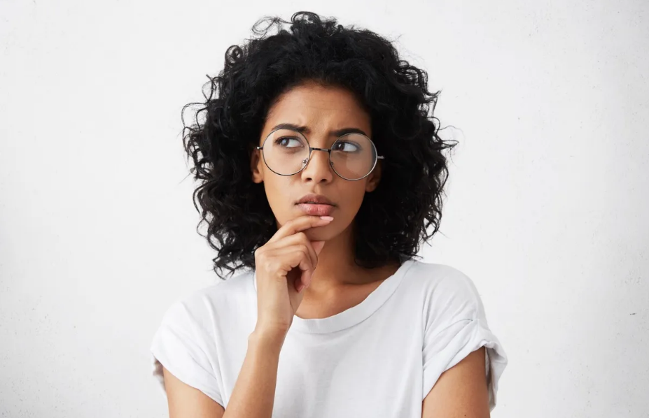 A pensive young woman with curly hair wearing round glasses and a white T-shirt, resting her chin on her hand against a white background.