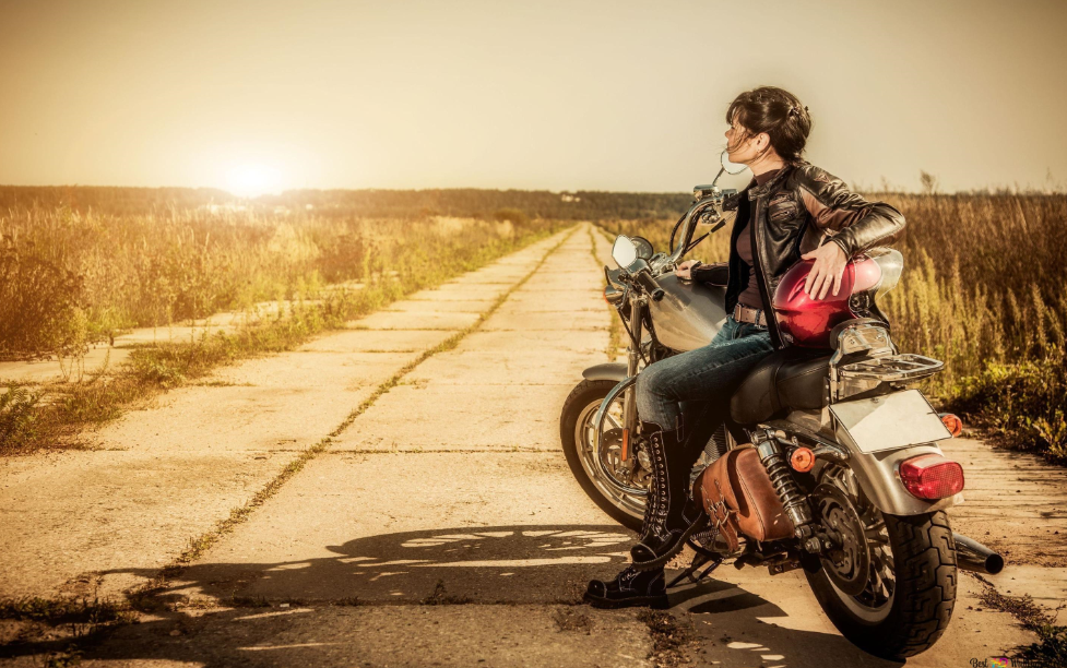 A person wearing a leather jacket and jeans sits sideways on a motorcycle parked on a rural road, facing the sunset with their head turned towards the sun.