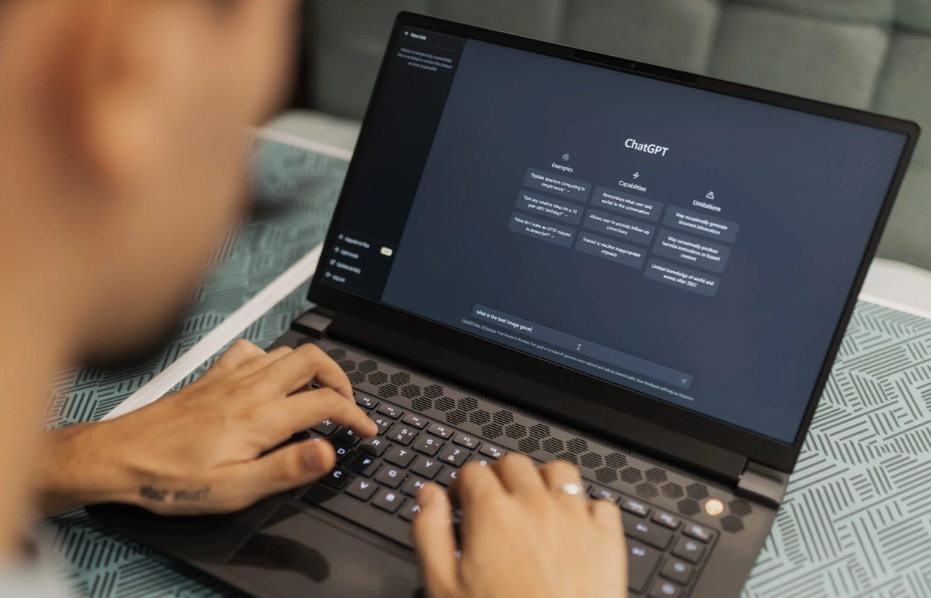 A person using a laptop showing a screen with "ChatGPT" interface, with options like "Examples," "Try it yourself," "Capabilities," and "Limitations."