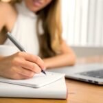 Person writing notes at desk with laptop and coffee mug for productive work session.