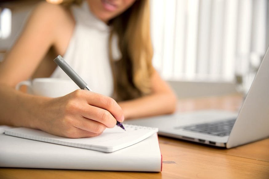 Person writing notes at desk with laptop and coffee mug for productive work session.