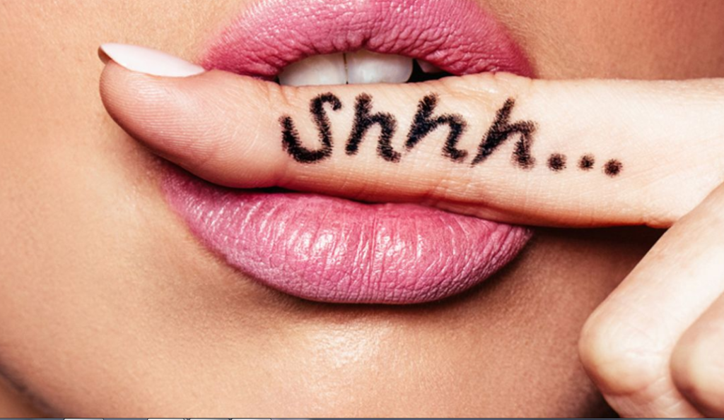 Close-up of a woman's pink lips with the word "shhh..." written on her index finger placed against them.