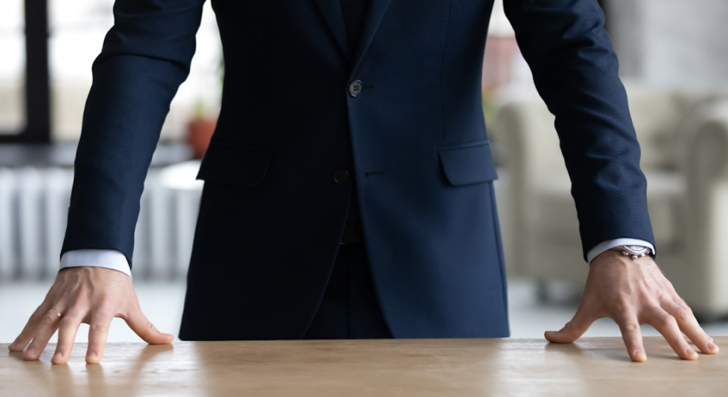 A person in a dark blue suit leaning over a table with hands firmly placed on the surface, a watch visible on the left wrist.