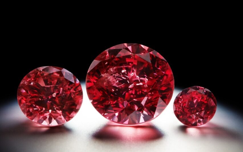 Three red gemstones of varying sizes displayed against a dark background with a prominent light source reflecting off their faceted surfaces.