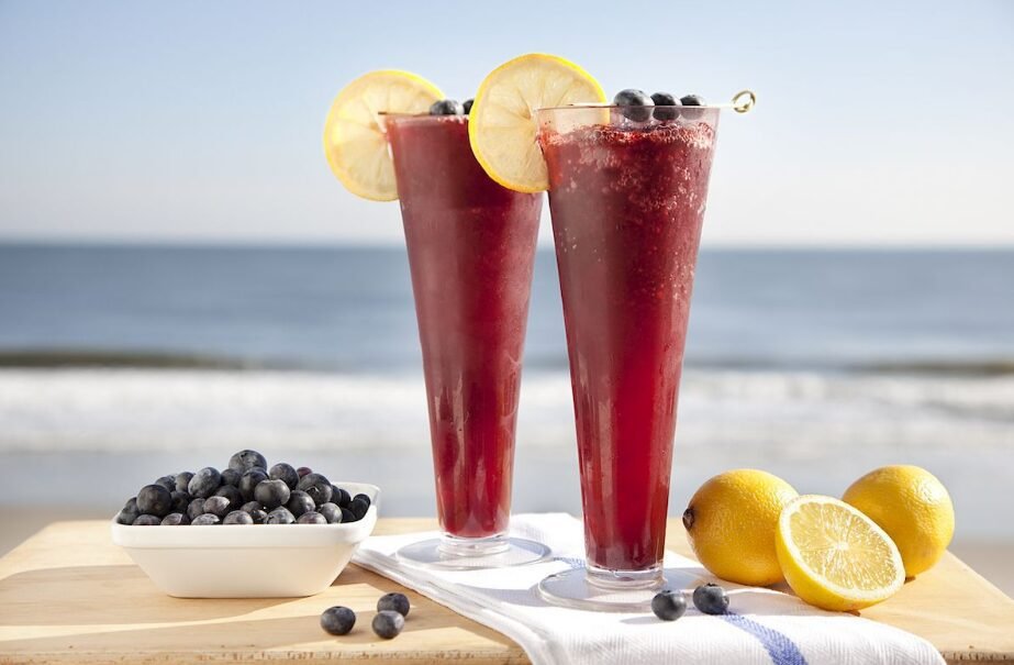 Two glasses of red smoothies garnished with lemon slices and blueberries, with a bowl of blueberries and halved lemons on a wooden surface, set against a beach backdrop.