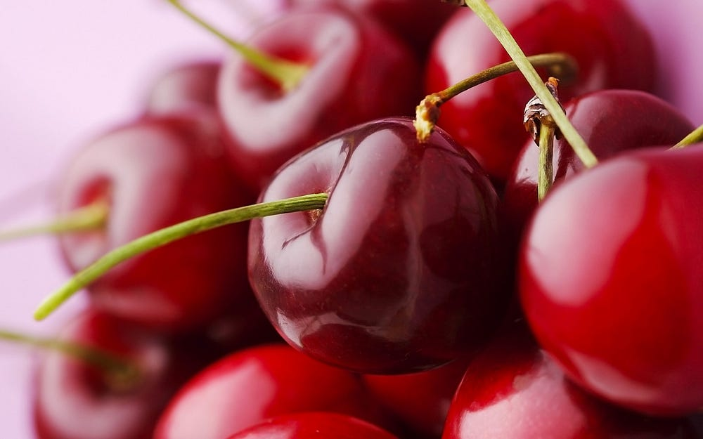 A close-up of a pile of glossy red cherries with green stems isolated on a pink background.