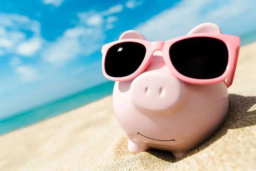 A pink piggy bank wearing pink sunglasses sits on a sandy beach with a clear blue sky in the background.